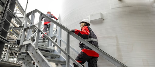 Worker in red and black safety clothing inspecting industrial equipment beside a large storage tank.