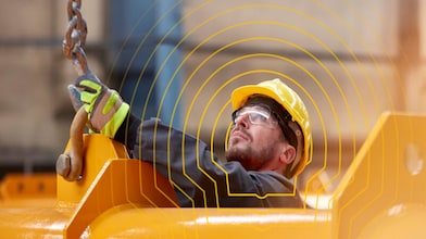 A worker wearing a yellow hard hat and safety glasses operating lifting equipment in an industrial setting.