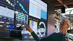 Man pointing at a large digital display filled with graphs, charts, and analytics inside a modern tech workspace.