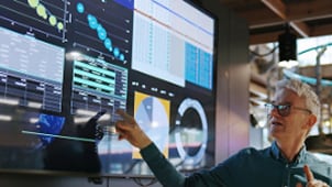 Man pointing at a large digital display filled with graphs, charts, and analytics inside a modern tech workspace.