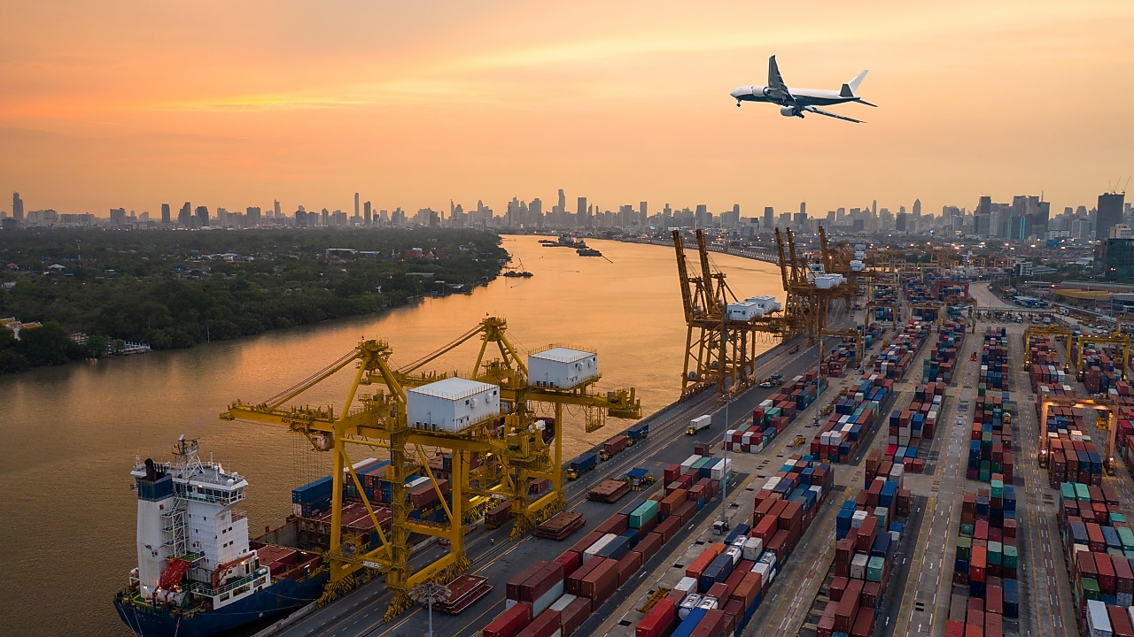 A large cargo port at sunset with stacked shipping containers, yellow cranes loading a cargo ship, and a commercial airplane flying overhead. A city skyline with high-rise buildings is visible in the background along a wide river.