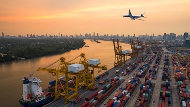 A large cargo port at sunset with stacked shipping containers, yellow cranes loading a cargo ship, and a commercial airplane flying overhead. A city skyline with high-rise buildings is visible in the background along a wide river.