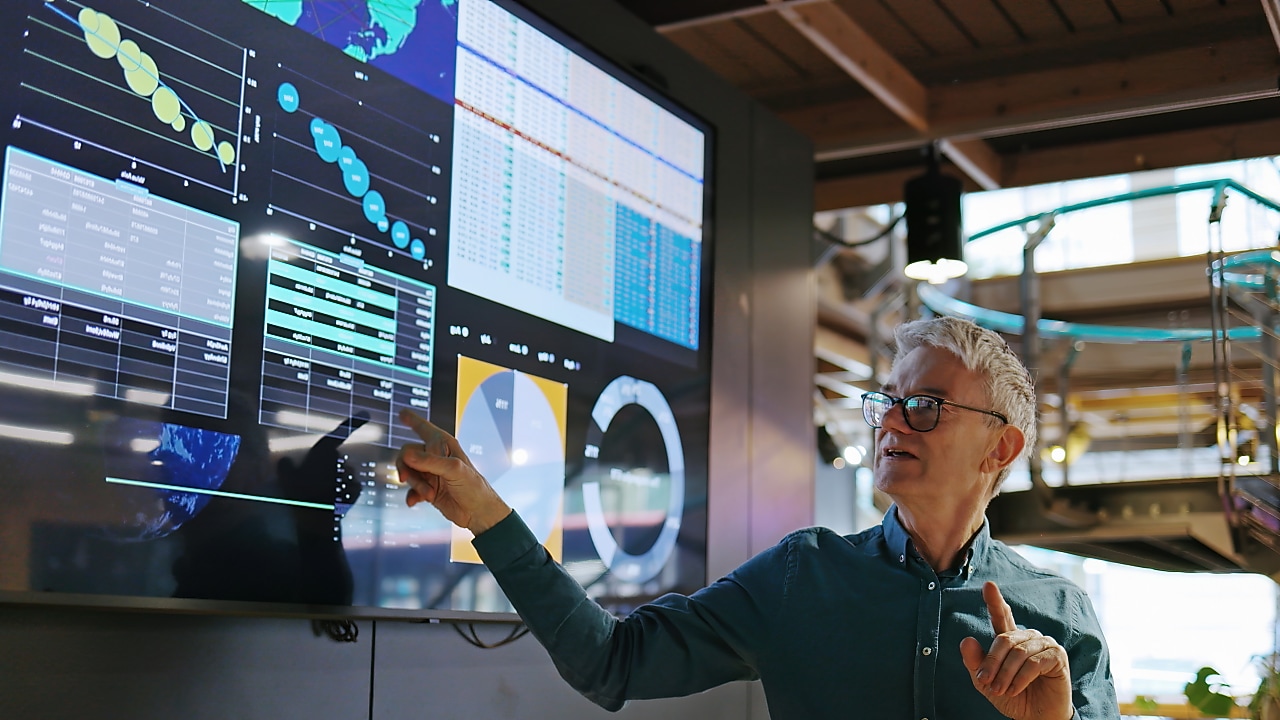 A man with grey hair and glasses, wearing a teal shirt, points at a large digital display screen showing various data visualizations, including charts, graphs, and maps.