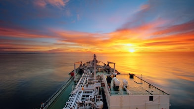 View from the deck of a large cargo or tanker ship at sea, heading into a vibrant sunset with calm waters and a dramatic sky.