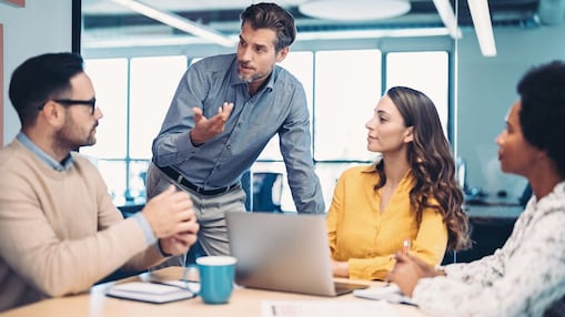 Four colleagues gathered around a table in a modern office, with one person standing and gesturing while discussing ideas with the seated team members around a laptop.