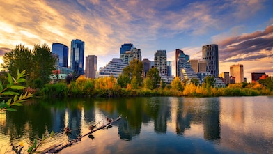 City skyline at sunset reflected in a calm lake, with modern high-rise buildings rising behind a line of autumn-colored trees and dramatic clouds overhead.
