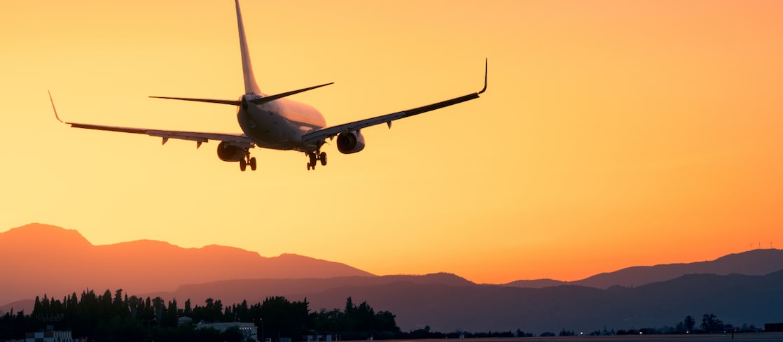 A commercial airplane coming in for landing against a vibrant orange sunset sky, with silhouettes of mountains and trees in the background.