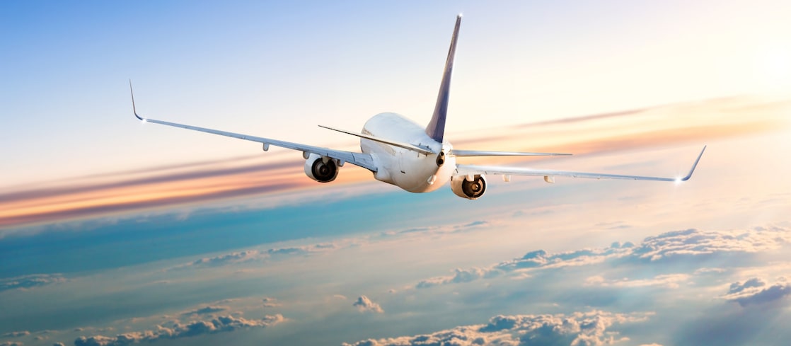 A commercial airplane flying at high altitude above the clouds during sunrise or sunset, with its tail and wings illuminated by the soft light.