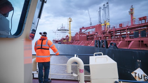 Worker in a high-visibility jacket standing on a tugboat, guiding a large red cargo ship in a busy industrial port.