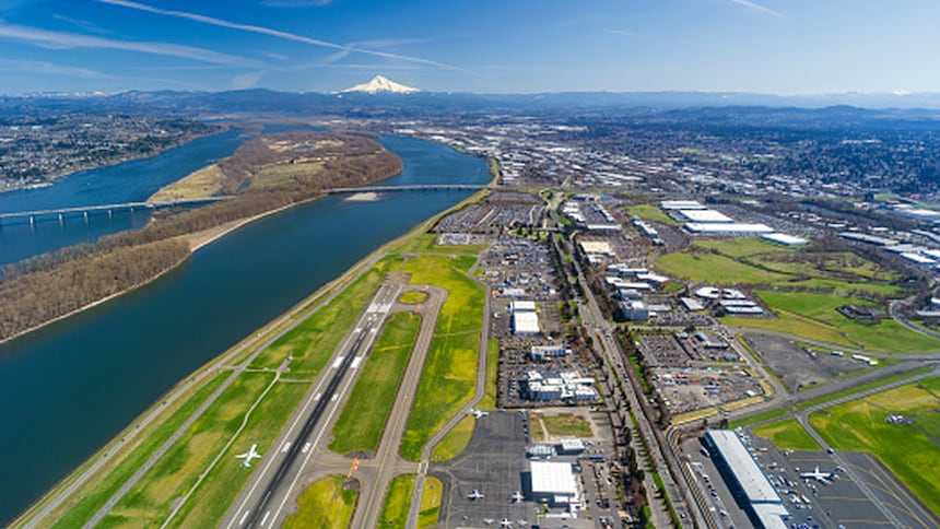 Aerial view of an airport runway beside a river, with surrounding industrial areas and mountains in the distance.