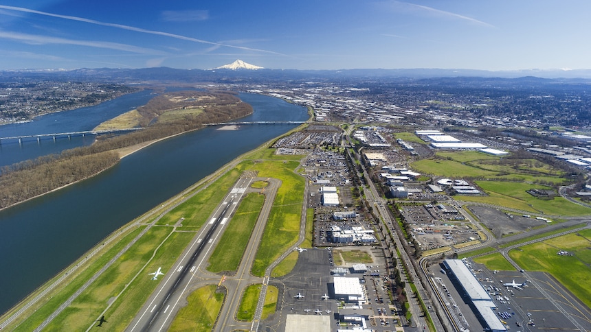 Aerial view of an airport runway beside a river, with surrounding industrial areas and mountains in the distance.