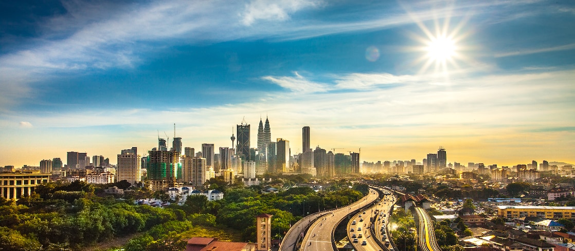 Wide view of a modern city skyline under a bright sun, with high-rise buildings in the distance and a busy highway curving through green areas in the foreground.