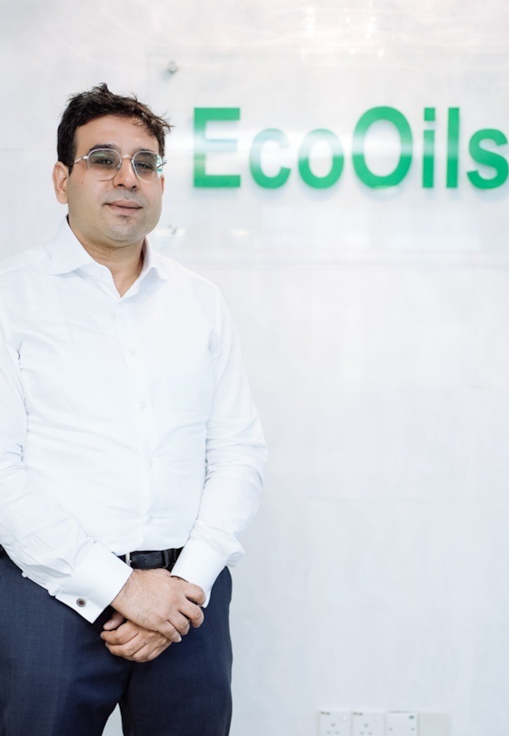 Rida stands in a corporate office setting in front of an EcoOils sign, wearing business attire and facing the camera.