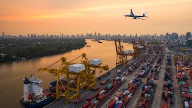 A large cargo port at sunset with stacked shipping containers, yellow cranes loading a cargo ship, and a commercial airplane flying overhead. A city skyline with high-rise buildings is visible in the background along a wide river.