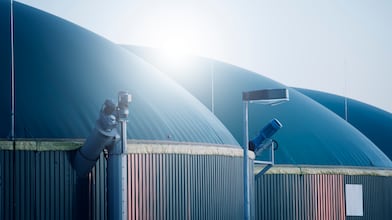 A large cargo port at sunset with stacked shipping containers, yellow cranes loading a cargo ship, and a commercial airplane flying overhead. A city skyline with high-rise buildings is visible in the background along a wide river.