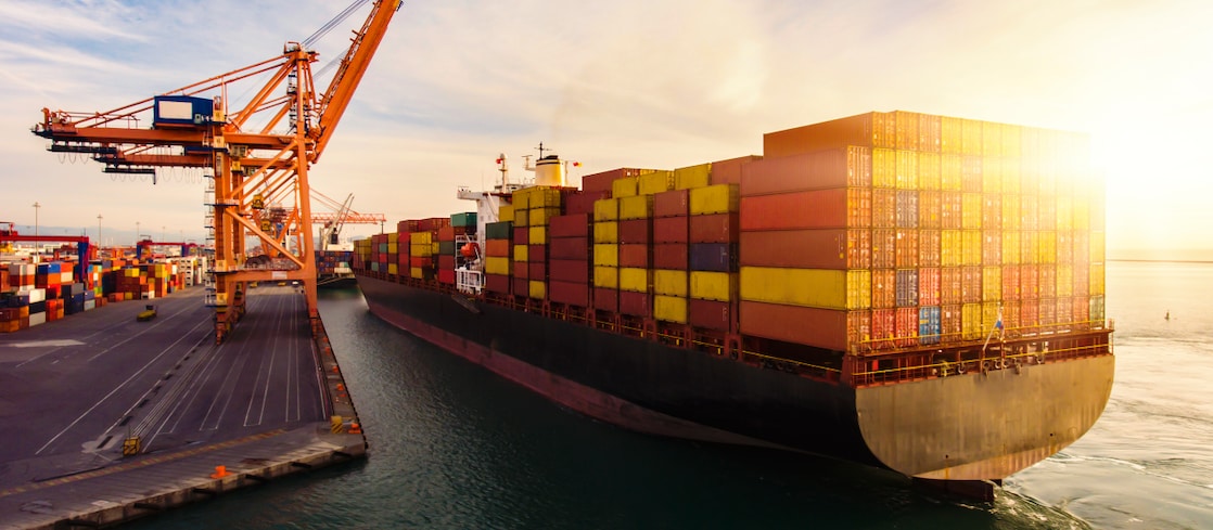 A large container ship loaded with multicoloured shipping containers docks at a port terminal, with cranes unloading cargo under a bright sunset sky.