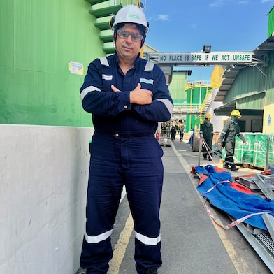 Rida stands at an industrial site wearing full PPE, including a helmet and safety gear, with equipment visible in the background.