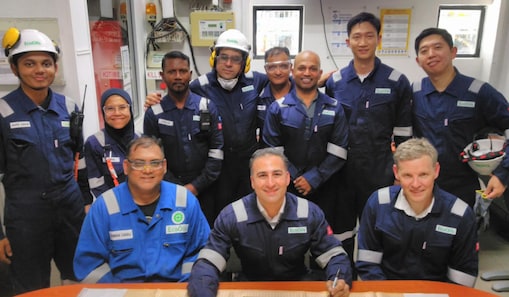Rida sits with a group of workers in a control room, posing together around a desk with equipment and documents.