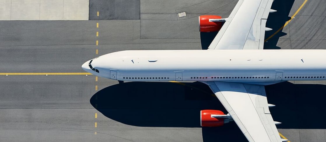 Top-down view of a commercial aircraft parked on an airport taxiway, with red engine nacelles and the aircraft's shadow visible on the tarmac.