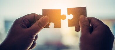 Two hands holding separate jigsaw puzzle pieces, positioned close together against a softly lit background, symbolising connection, collaboration, or problem solving.