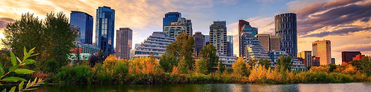 City skyline at sunset reflected in a calm lake, with modern high-rise buildings rising behind a line of autumn-colored trees and dramatic clouds overhead.