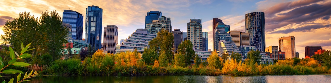 City skyline at sunset reflected in a calm lake, with modern high-rise buildings rising behind a line of autumn-colored trees and dramatic clouds overhead.