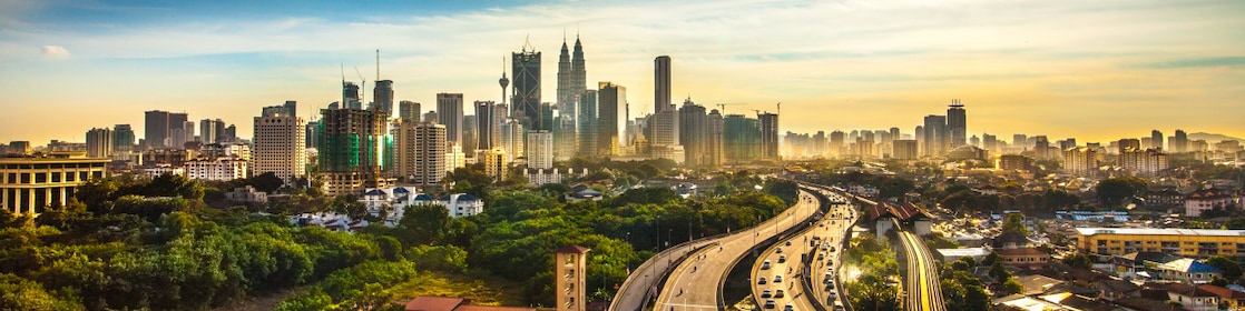Wide view of a modern city skyline under a bright sun, with high-rise buildings in the distance and a busy highway curving through green areas in the foreground.