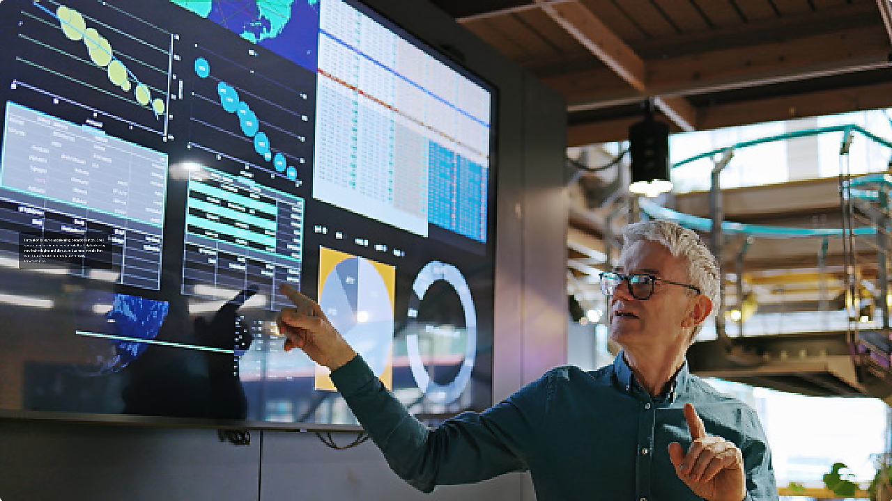 Man pointing at a large digital display filled with graphs, charts, and analytics inside a modern tech workspace.