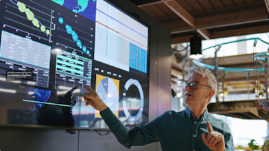 Man pointing at a large digital display filled with graphs, charts, and analytics inside a modern tech workspace.