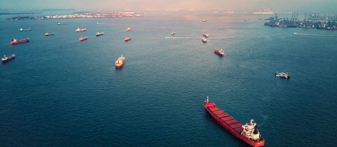 Aerial view of multiple cargo ships and tankers navigating a busy shipping channel near a port, with cranes and industrial facilities visible on the horizon under partly cloudy skies.