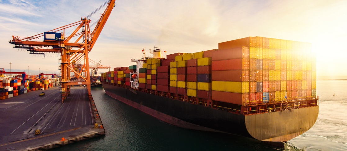 A large container ship loaded with multicoloured shipping containers docks at a port terminal, with cranes unloading cargo under a bright sunset sky.