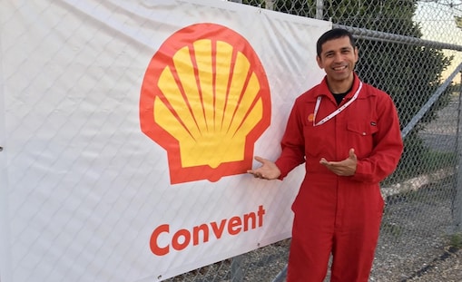 A Shell worker in a red coverall uniform gives a thumbs-up in front of a banner with the Shell logo and the word “Convent.”