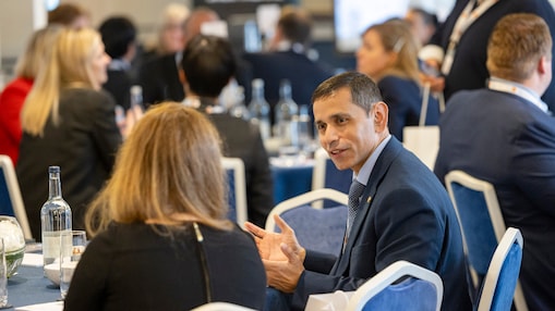Rakshay Shah, in a suit, talk to an attendee across a table at a professional networking or conference event.