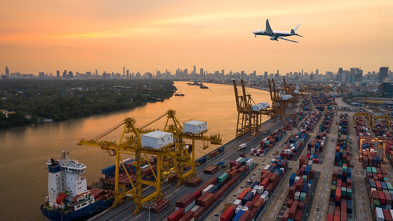 Aerial view of a busy container port with cranes and shipping containers, as a plane flies overhead during sunset with a city skyline in the background