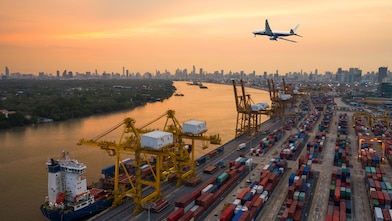 Aerial view of a busy container port with cranes and shipping containers, as a plane flies overhead during sunset with a city skyline in the background