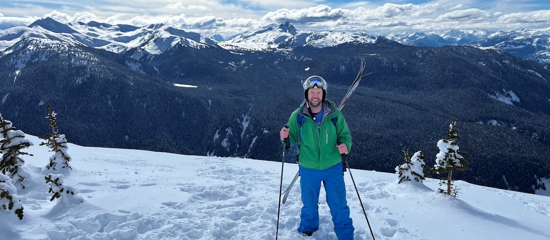 Hector MacQuarrie standing on a snowy mountain slope with skis and poles, smiling at the camera. He’s wearing a green jacket, blue pants, and a helmet with goggles.