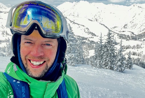 Hector MacQuarrie taking a selfie on a snowy mountain slope. He’s wearing a green jacket, blue gear, and reflective ski goggles, with snow-covered pine trees and bright white mountain peaks stretching into the distance behind him under a partly cloudy sky.