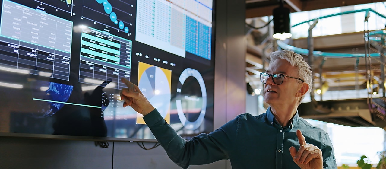 An engineer or data analyst presenting information on a large digital screen displaying charts, graphs, and data visualizations inside a modern industrial or research facility.