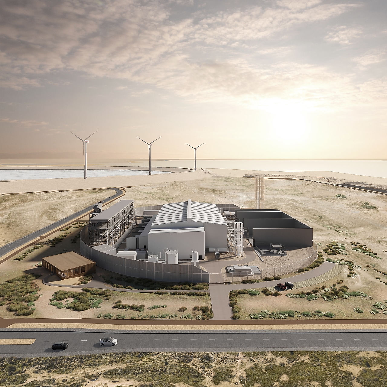 An aerial view of Shell’s Holland Hydrogen 1 facility set in a desert landscape near the coast, featuring large metallic buildings, storage tanks, and surrounding infrastructure, with wind turbines visible in the background under a setting sun.