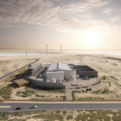 An aerial view of Shell’s Holland Hydrogen 1 facility set in a desert landscape near the coast, featuring large metallic buildings, storage tanks, and surrounding infrastructure, with wind turbines visible in the background under a setting sun.