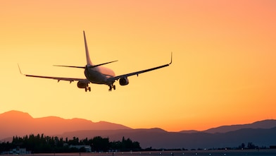 A commercial airplane coming in for landing against a vibrant orange sunset sky, with silhouettes of mountains and trees in the background.
