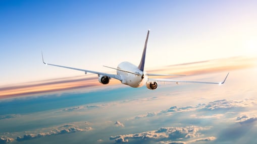 A commercial airplane flying at high altitude above the clouds during sunrise or sunset, with its tail and wings illuminated by the soft light.