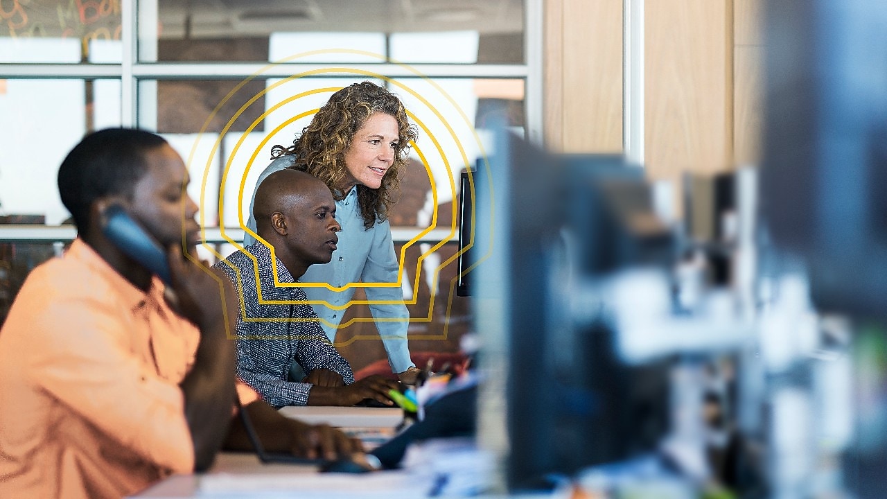Three people collaborating at computer workstations in a modern office with large windows and multiple monitors.