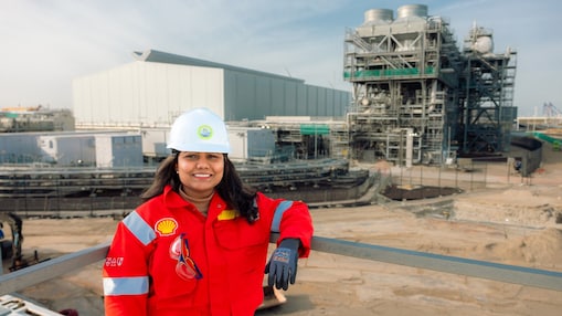 A person wearing a red Shell protective suit and a white safety helmet stands outdoors on an industrial site, with gloved hand resting on a railing