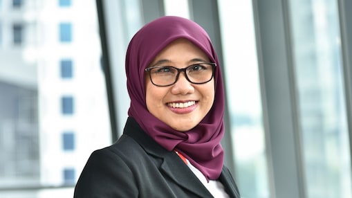 A young woman smiling, wearing a purple hijab, a dark jacket, and glasses, against office background.