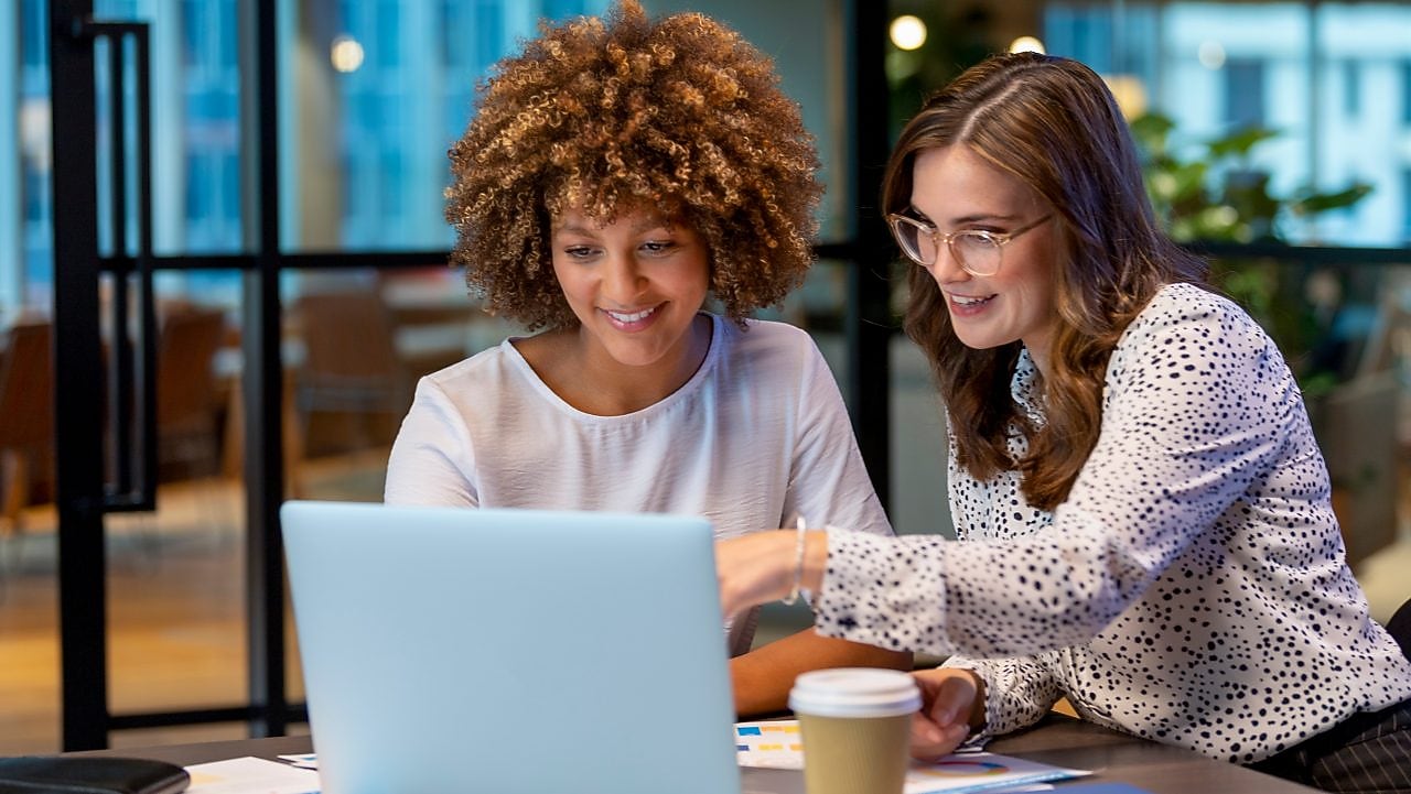 Two professionals collaborating at a table with a laptop, documents, and a coffee cup in a modern office setting, with large windows and contemporary furniture in the background.