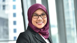 A young woman smiling, wearing a purple hijab, a dark jacket, and glasses, against office background.