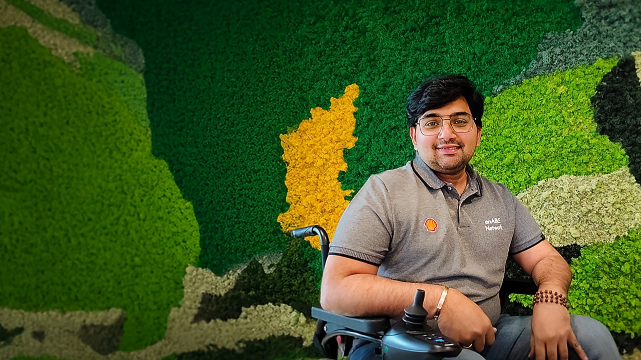  Person in a wheelchair wearing a Shell enABLE Network shirt, seated against a green and yellow moss wall.