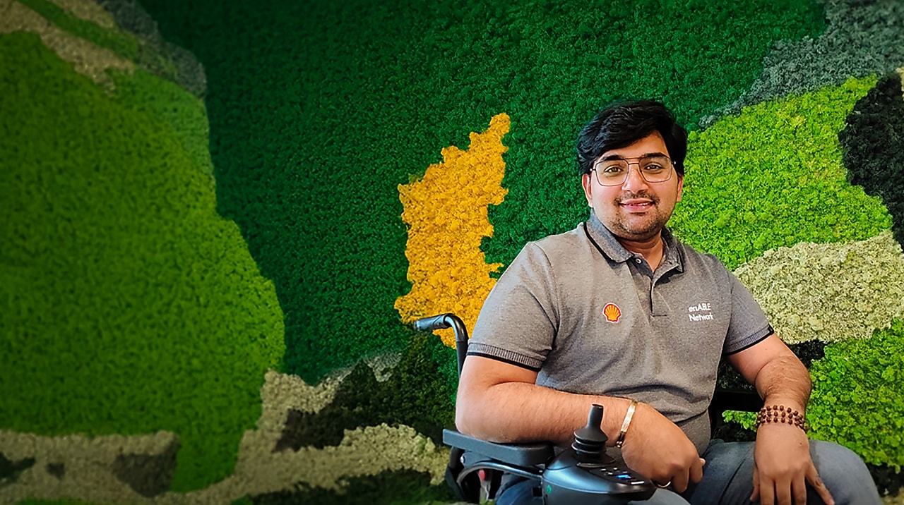  Person in a wheelchair wearing a Shell enABLE Network shirt, seated against a green and yellow moss wall.
