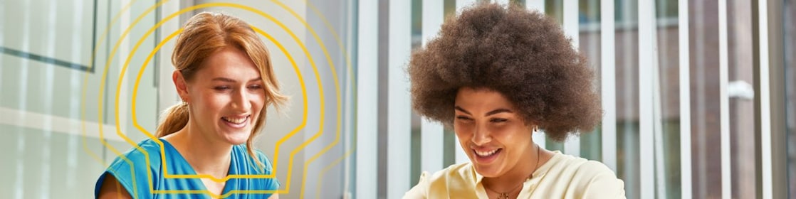 Two women sitting at a table working on laptops in a bright office with vertical blinds and a coffee cup nearby